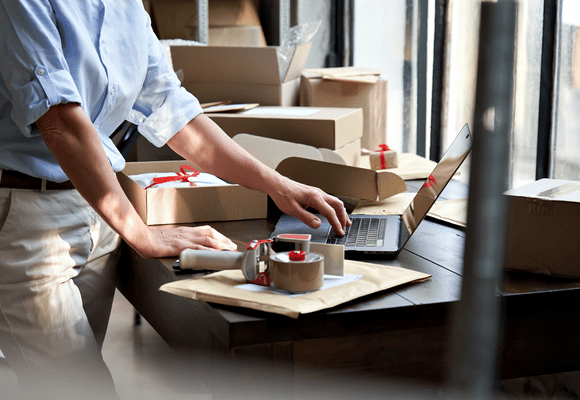 a side view of a business owner working on inventory on their laptop with shipping boxes around them