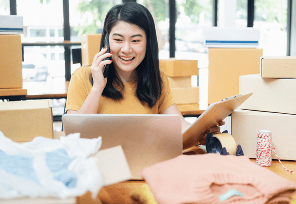 a smiling female business owner working on her laptop while talking on the phone, surrounded by boxes 