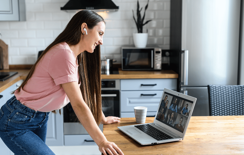 a photo of a girl inside of her kitchen leaned over her counter having a group zoom call on a laptop